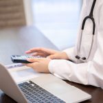 Doctor in a white coat using a calculator next to a laptop on a desk.